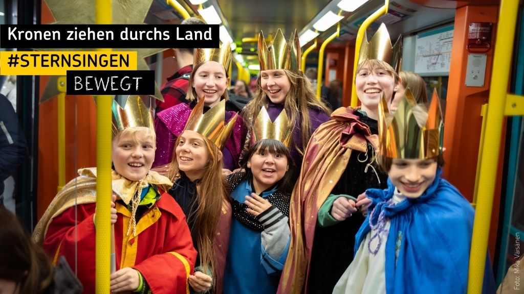 Sieben Kinder in Königsgewändern mit Kronen stehen lachend in einer Straßenbahn; links oben Text: 'Kronen ziehen durchs Land, #Sternsingen bewegt'