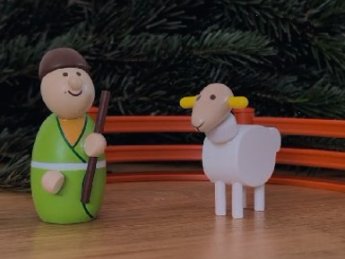 Hirte und Schaf aus Holz, angemalt stehen vor einem kleinen Zaun, im Hintergrund Tannen
