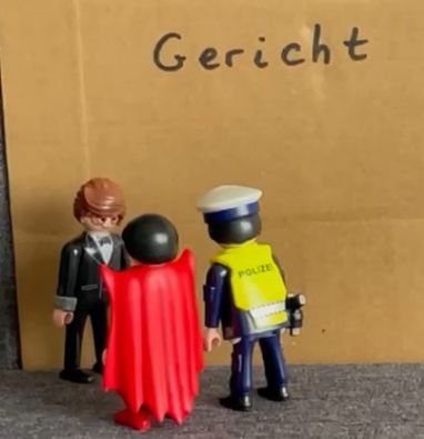 Pappe mit dem Wort 'Gericht' davor zwei Playmobilmännchen , die einen dritten befragen.