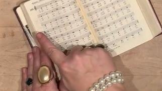 offenes Gebetbuch, eine Frauenhand, die mit Ringen und einem Perlen-Armbband reich geschmückt ist.
