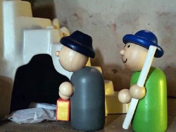 Zwei Holzmännchen stehen vor einem offenen Höhlengrab. Am Boden liegt ein weißes Tuch.